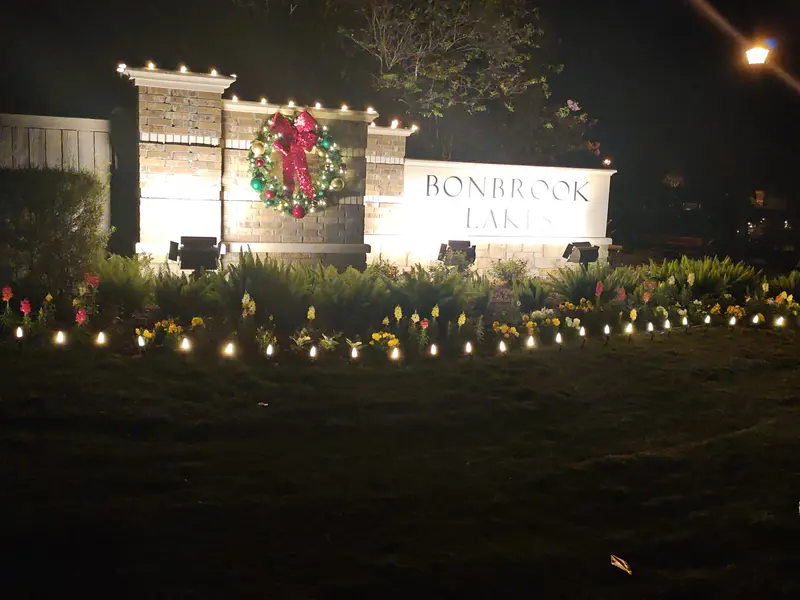 Community entrance monument lettered Bonbrook Lakes at night, with a decorated wreath and red bow on the brick pier, warm-white bulbs along the top of the wall and a staked run of bulbs along the flower bed in front