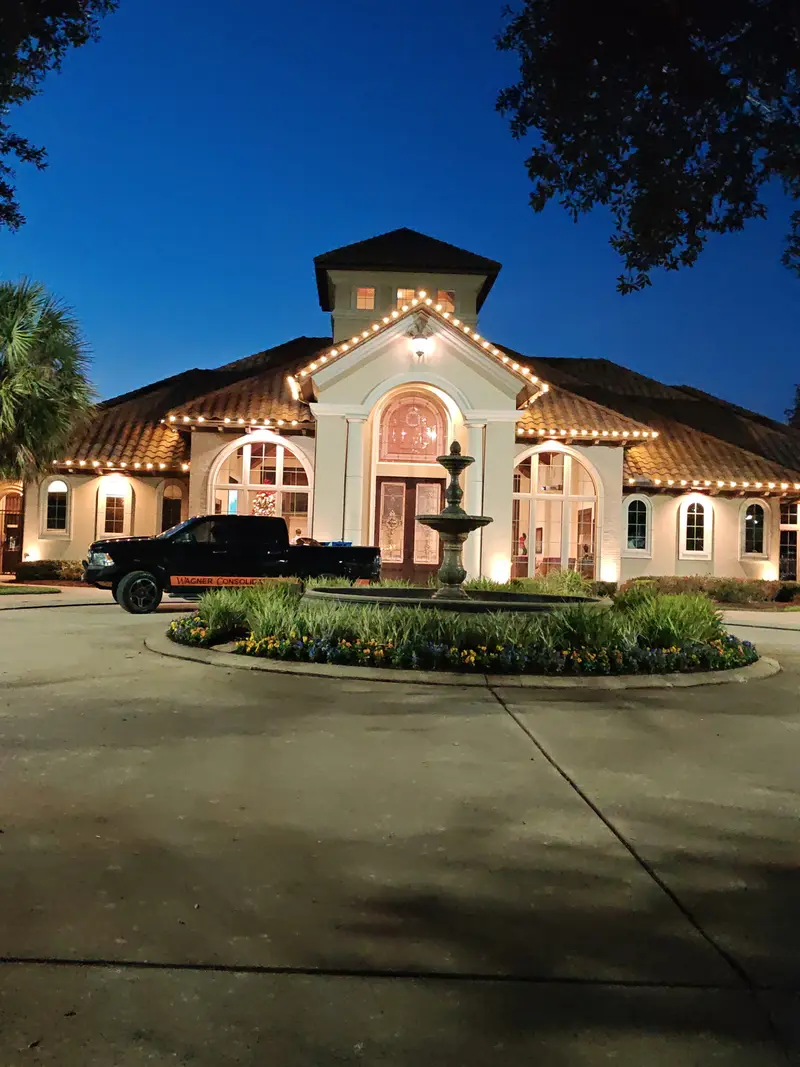 Stucco clubhouse at dusk with warm-white bulbs following the tile roofline and gables, a stone fountain in the circular drive and a pickup truck lettered Wagner Consolidated parked at the entrance