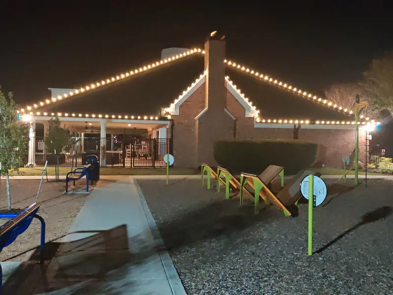 Single-storey brick clubhouse at night with warm-white bulbs following each roofline and the chimney gable, seen across the playground equipment in front of it