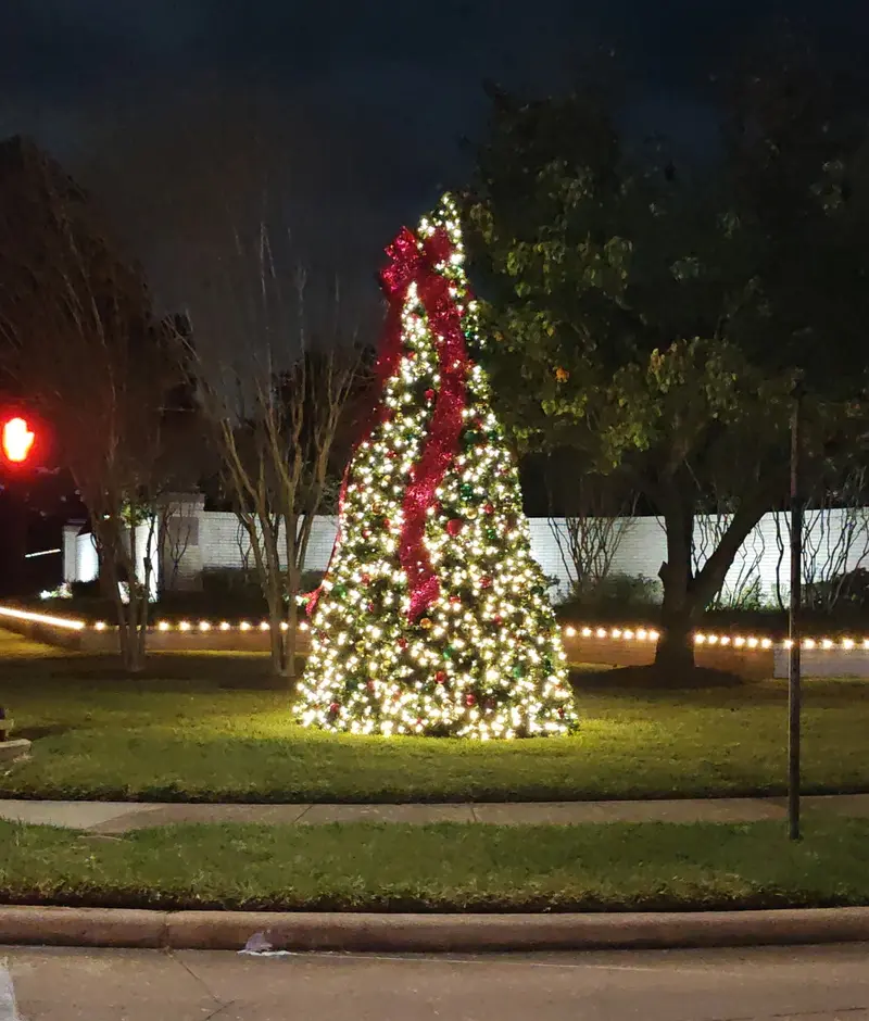 Tall Christmas tree lit in warm white on a grass median at an intersection, with a wide red ribbon spiralling down it, hanging ornaments and a run of warm-white bulbs along the border behind