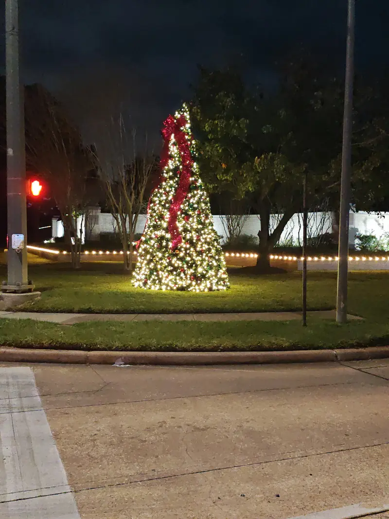 Tall Christmas tree lit in warm white on a grass median at an intersection, with a wide red ribbon spiralling down it, hanging ornaments and a run of warm-white bulbs along the border behind
