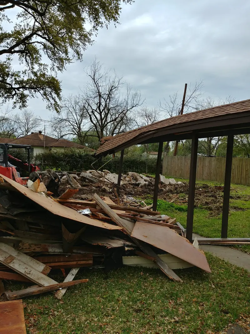 Pile of demolished lumber and sheet goods on a residential lawn, with the arm of a loader visible at the left edge and a dirt patch under a carport behind it