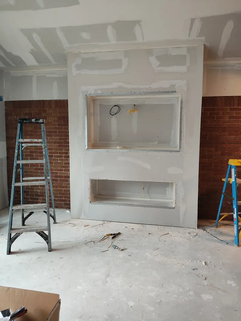 Taped drywall wall with two open niches, a step ladder on the left and a brick return on each side