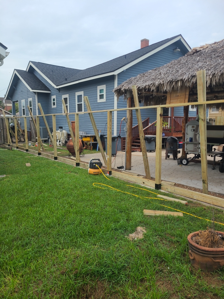 New wood fence posts and a bottom rail across a backyard, panels not yet hung, a thatched roof and a smoker in the background