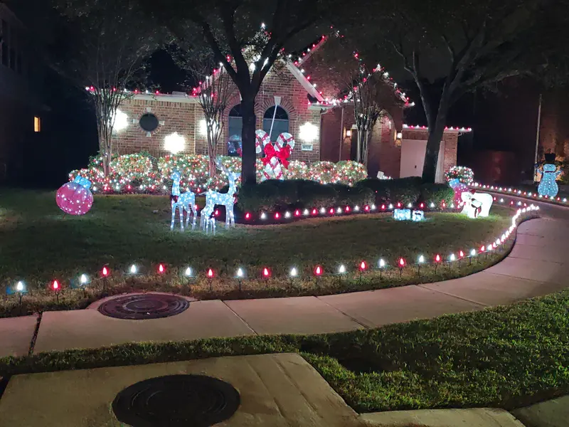 Brick house at night with bulbs along the rooflines and porch, hedges covered in red, white and green lights, lit reindeer, a snowman and gift boxes on the lawn, and red and white bulbs staked along the curving front path