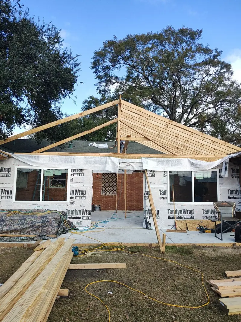 House wrapped in white house-wrap with open timber roof framing and lumber on the grass