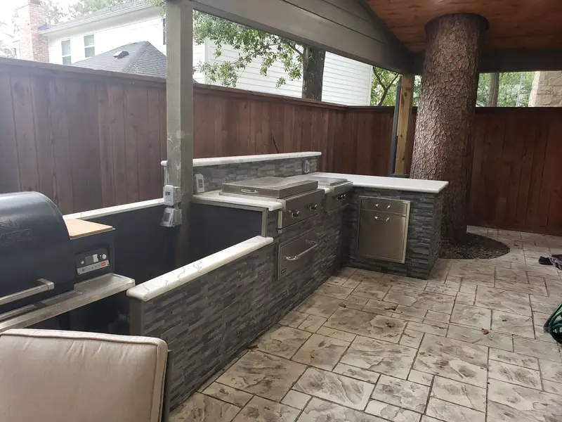 L-shaped outdoor kitchen with a stone face, white counter, stainless grill and a tree trunk rising through the patio