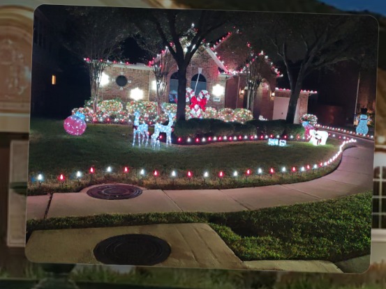 Suburban brick house at night with red and white Christmas lights along the rooflines, lit bushes, wrapped trees, illuminated yard figures including reindeer and a snowman, and red and white stake lights along the front walk