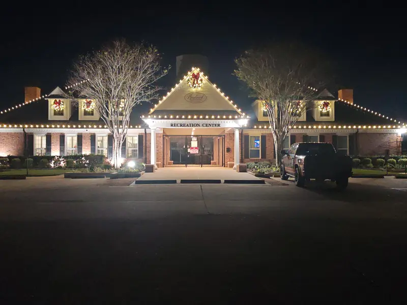 Two-storey brick recreation center at night with warm-white bulbs along every eave and gable, a lit wreath with a red bow on the centre gable and further lit wreaths in the dormer and ground-floor windows