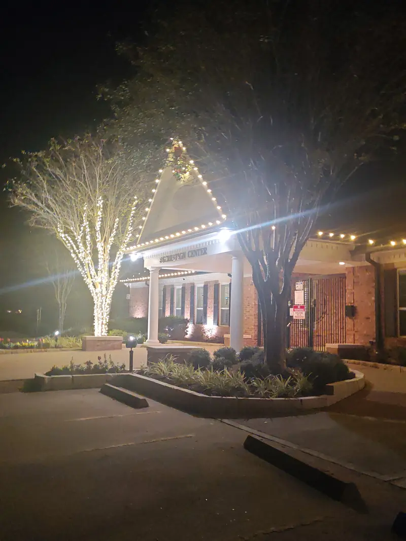 Recreation center entrance at night with warm-white bulbs along the gable and porch beam, and a bare tree beside it wrapped trunk to branch in the same warm-white lights